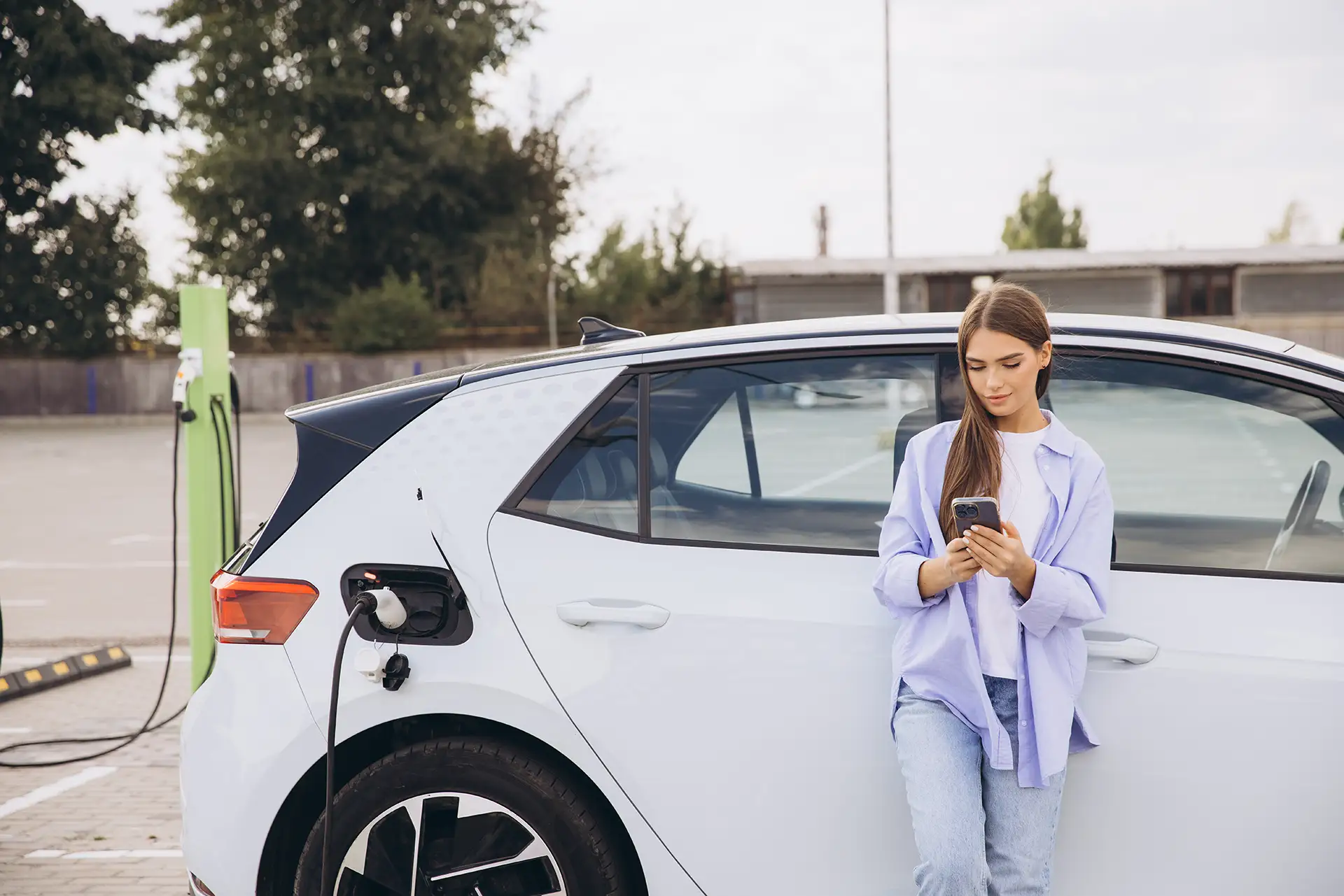 Junge Frau genießt die Zeit an einer Ladestation für Elektrofahrzeuge und nutzt dabei ihr Smartphone.